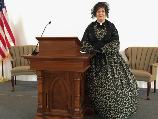 A woman wearing a long, black and yellow flowered dress, a black lace shawl, and a black hat and leaning on a podium.