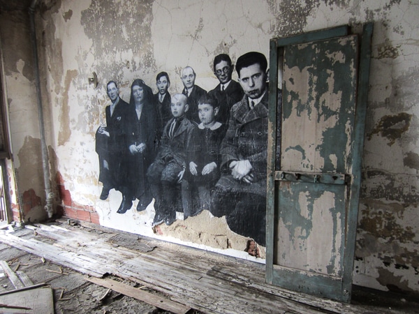 A life-sized black-and-white photo plastered to a wall in an extremely dilapidated room. The photo features five adults and two children. 