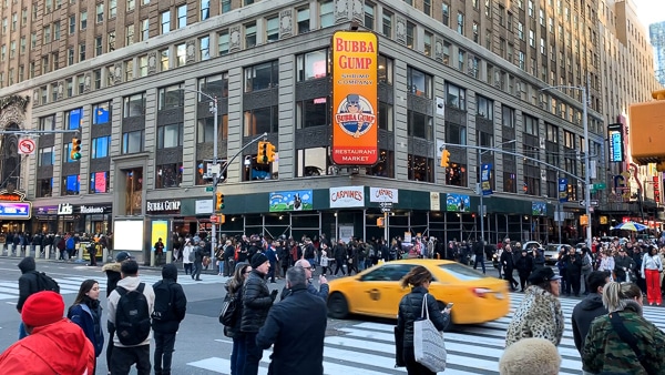 An intersection in Times Square with a lot of people crossing the streets. Bubba Gump and Carmines Restaurant are on the corner. 