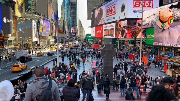 Standing near the top of the Red Steps in Times Square, looking down the steps. Crowds, billboards, and traffic are spread throughout the photo. 