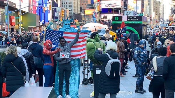 A tourist is holding a sign that says I "heart" NY. He is standing in front of a person wearing a Statue of Liberty costume, and next to a person dressed as Spiderman. A woman is taking their photo. 