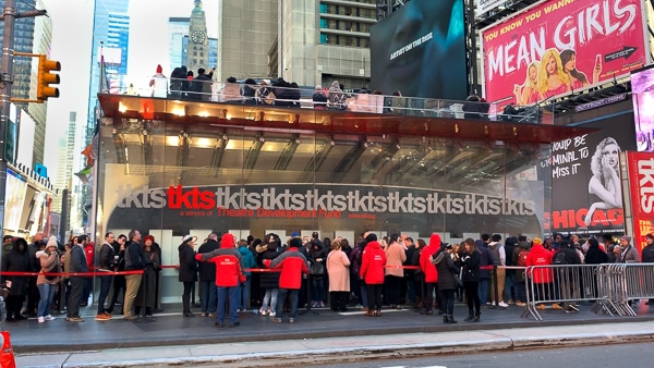 TKTS Discount Booth in Times Square. Several people are in line. The Red Steps are on top of the booth. 