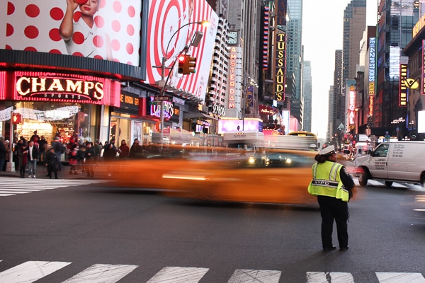 A Times Square intersection. A police officer wearing a neon yellow vest is directing traffic. Two taxis speed by in a blur. Champs store is on the corner of the intersection. 