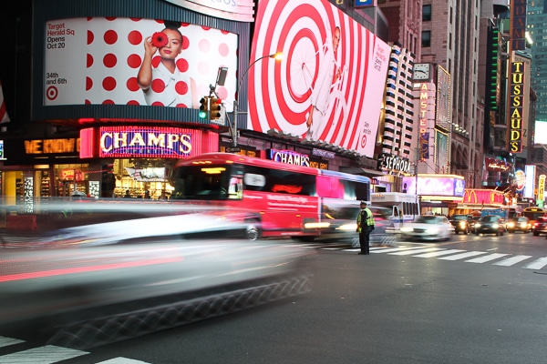 A Times Square intersection. A police officer wearing a neon yellow vest is directing traffic. A red bus and a car speed by in a blur. Champs store is on the corner of the intersection. 