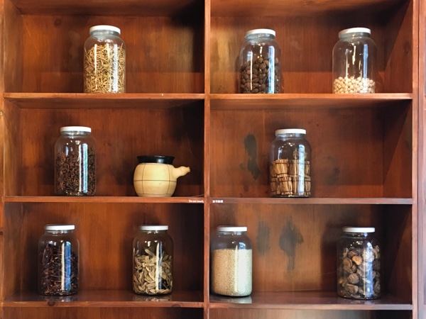 A wall shelf with six compartments. On each shelf sits a large jar of dried goods that appear to be herbs. 