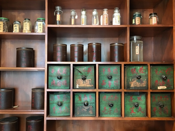 A brown wooden shelf showing about nine compartments. On the shelf sits bottles and tins of various sizes, and two rows of green tin boxes with red writing in Chinese. 