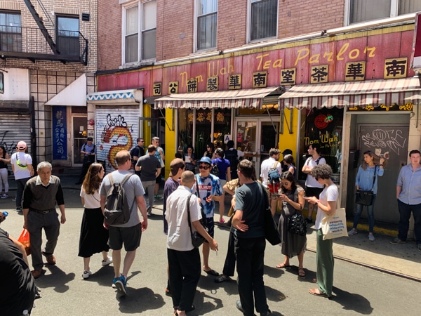 Crowd of people on Doyers Street in Chinatown; standing in front of Nom Wah Tea Parlor. The restaurant's sign is a faded pink with yellow writing. Chinese writing in black sits below the English name of the restaurant. 