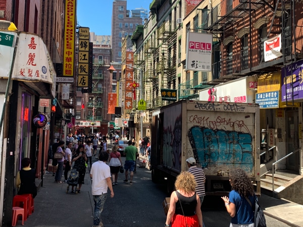 A narrow street in Chinatown on a warm summer day. Signs written in Chinese line the buildings, many people are walking, and a delivery truck is parked in front of a hair salon.