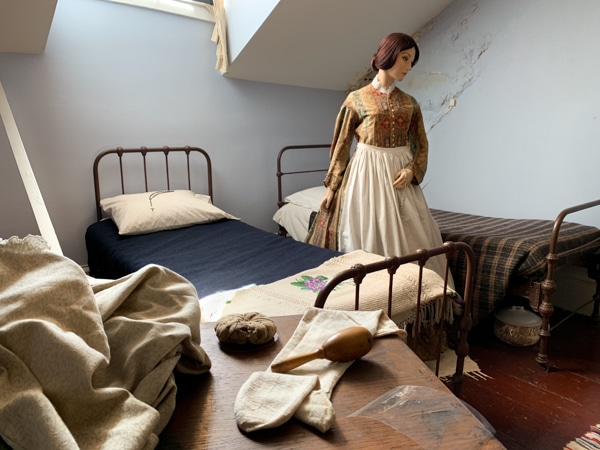 Two twin beds with iron frames. IN between the beds, a female mannequin is dressed to look like a servant. 