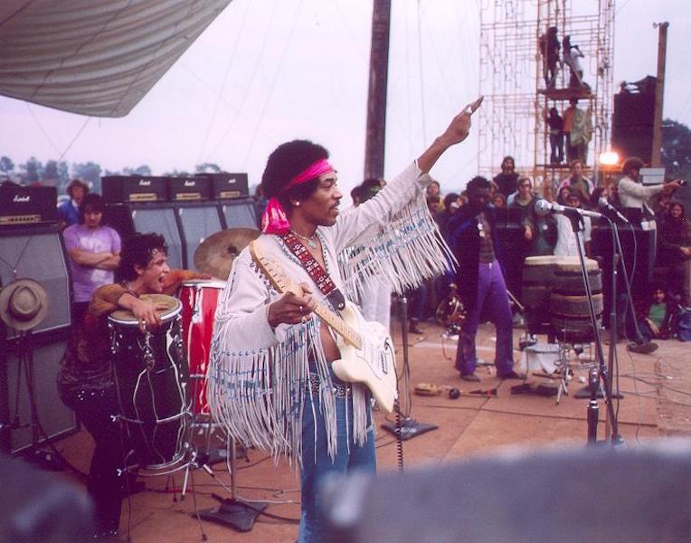 Jimi Hendrix on stage at Woodstock 1969. He is wearing a white fringed jacket and a red headband and holding a white guitar.