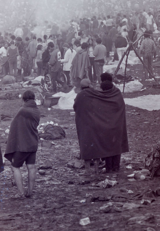 Black-and-white photo of Woodstock 1969 festival. In the foreground, people are wrapped in blankets. In the background, a large crowd is milling around. The shot is from behind, so mostly people's backs can be seen. Garbage is scattered on the ground.