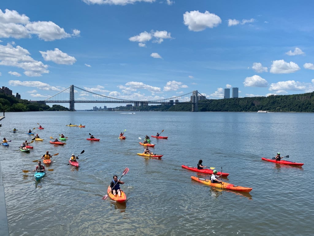 About a dozen people kayaking on the Hudson River. The George Washington Bridge is in the background.