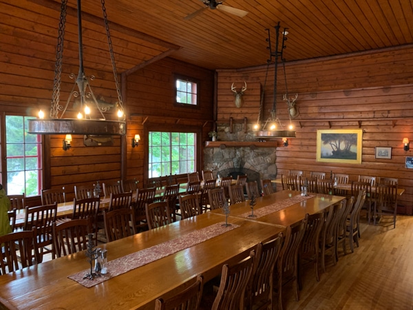 Dining hall at Great Camp Sagamore. Interior is wood paneled. Two low chandeliers hang over a long dining table. 