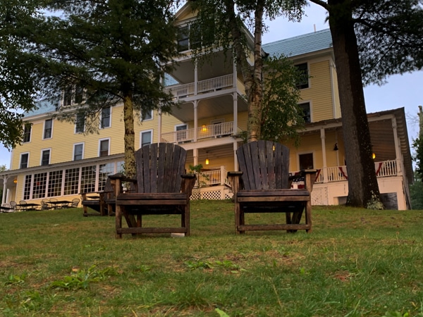 Exterior of The Woods Inn; the inn is a four-story, yellow building. Two Adirondack chairs sit in the foreground. 