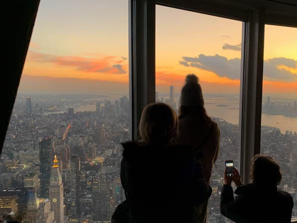 Three people seen from behind, looking out of floor-to-ceiling windows out onto the view of New York City. 