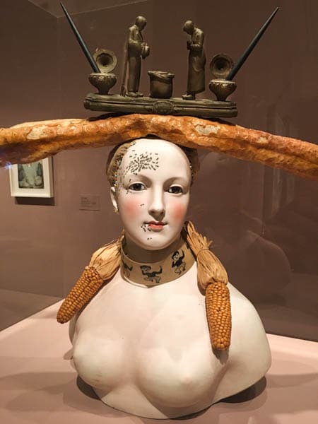 Porcelain bust of a woman with a baguette and an inkwell on her head, two ears of corn wrapped around her neck like a scarf, and painted ants crawling on her face.