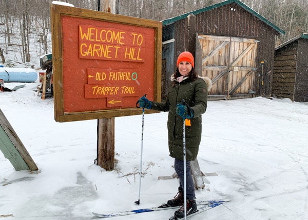 A woman wearing cross country-skis, standing  in front of a large orange wooden sign that says Welcome to Garnet Hill.