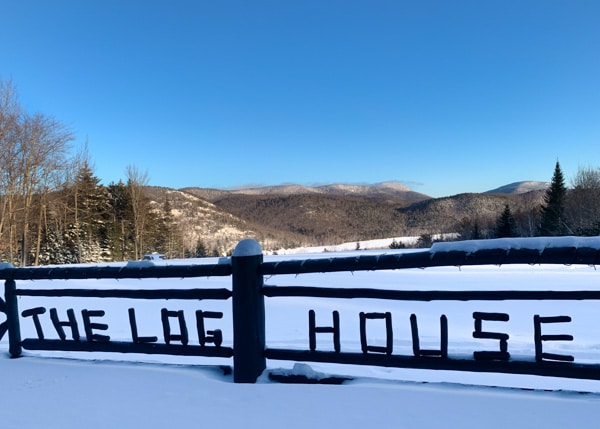A view from the deck at the Log House, looking out onto the mountains. The sky is very blue and the words, The Log House is spelled out in wood and embedded into the deck's side railing.
