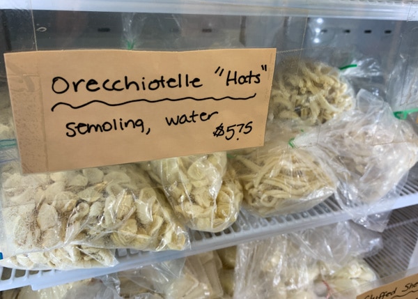 Several clear plastic bags full of homemade pasta inside a refrigerator. Handwritten sign says Orecchiotelle "Hats" Semolina, Water. $5.75.