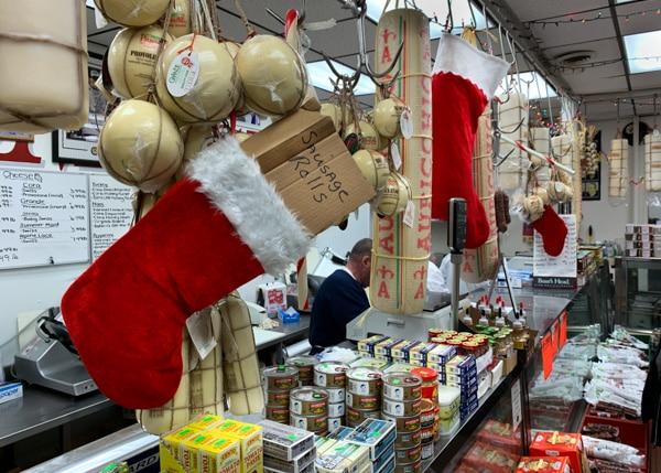 A deli counter stocked with  several packaged food items. In the foreground, a Christmas stocking hangs from the ceiling with a box labeled "Sausage Rolls" inside. Several balls of cheese also hang from the ceiling.