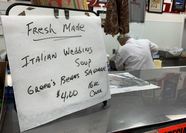 A handwritten sign on top of a deli counter. The sign says, "Fresh Made Italian Wedding Soup, Greens, Beans, Sausage $4.00, 16 oz. container"