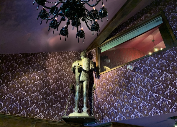 Motel room with a high ceiling and a black chandelier hanging from the ceiling. A green metal knight in armor hangs on the wall behind the chandelier. 