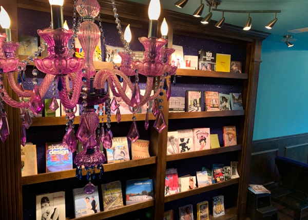 A pink chandelier hangs from the ceiling in the foreground with a floor-to-ceiling bookshelf behind it. 