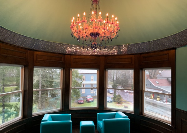Shimmer Spa. Two turquoise leather chairs and small, matching ottoman sit in front of a row of bay windows. An orange chandelier hangs from the ceiling above the chairs.