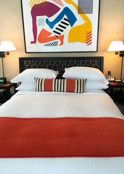 Wallcourt Hall guest room with queen-sized bed covered in a white duvet. An orange throw and orange and black striped pillow are on top of the bed. A colorful modern art painting hangs above the bed.