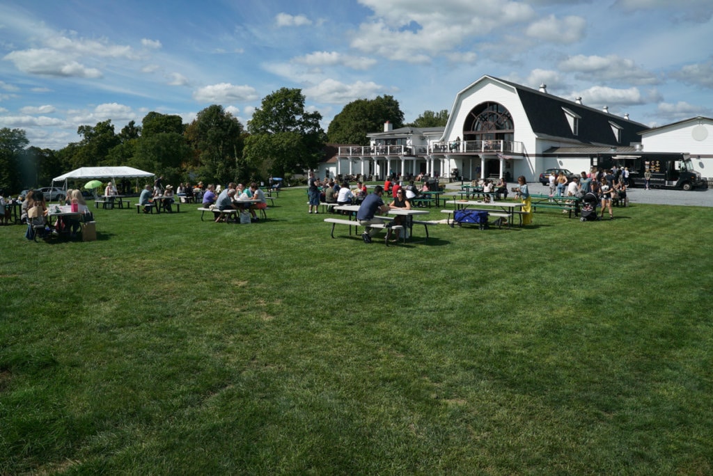 Several picnic tables on a large lawn. A huge white barn is in the background. 