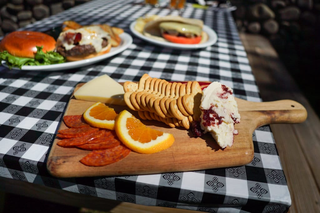 A wooden cheese board topped with salami, oranges, crackers, and cheese. It is sitting on a picnic table with a checkered black and white table cloth.
