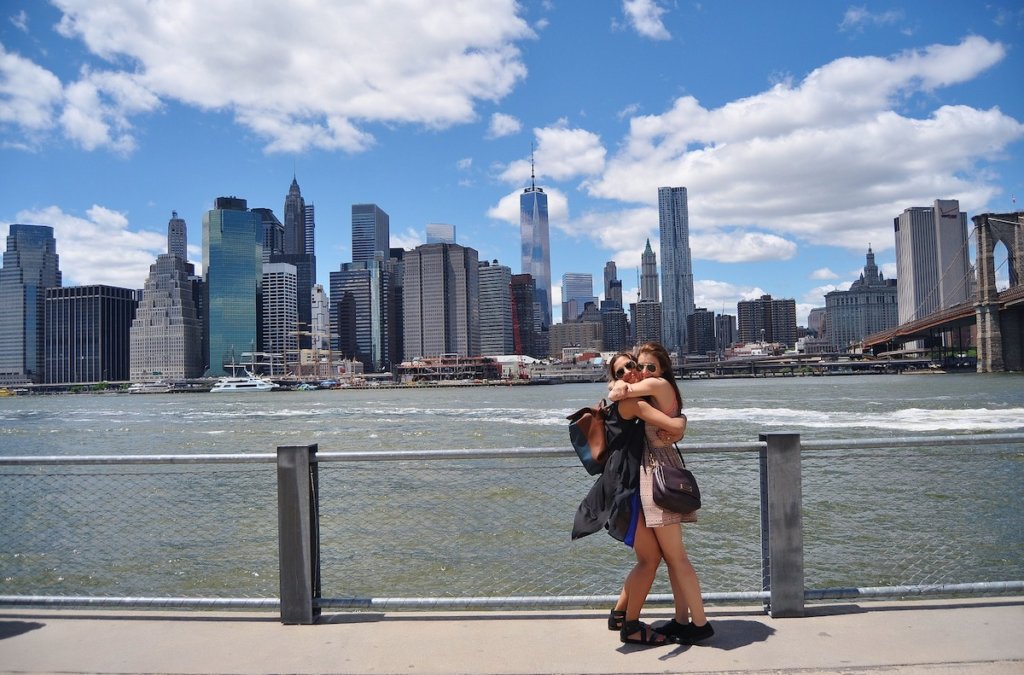 Two women hug each other with the skyline of New York City in the background. 