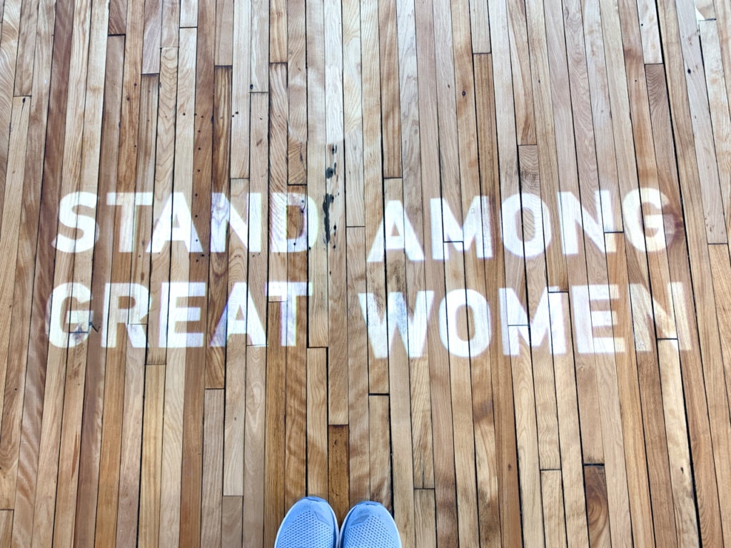 The phrase "Stand Among Great Women" projected onto a hardwood floor. 