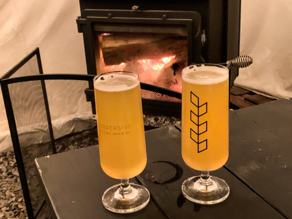 Two draft beers sitting on a table with a fire in a woodstove behind it.