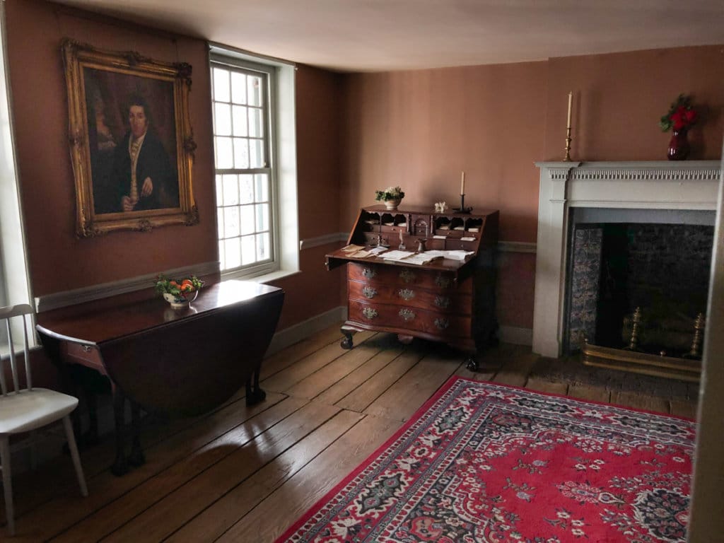 Parlor with colonial-era desk, table, and rug. Oil portrait hanging on wall. 