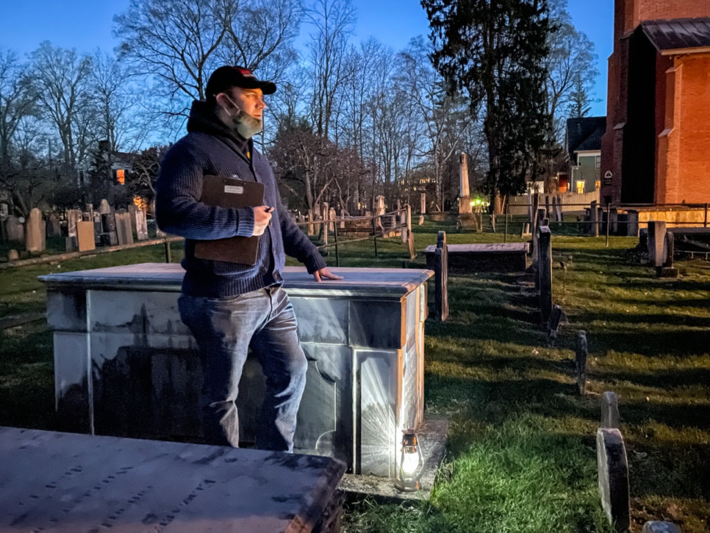 Tour guide stands in a graveyard at dusk. 
