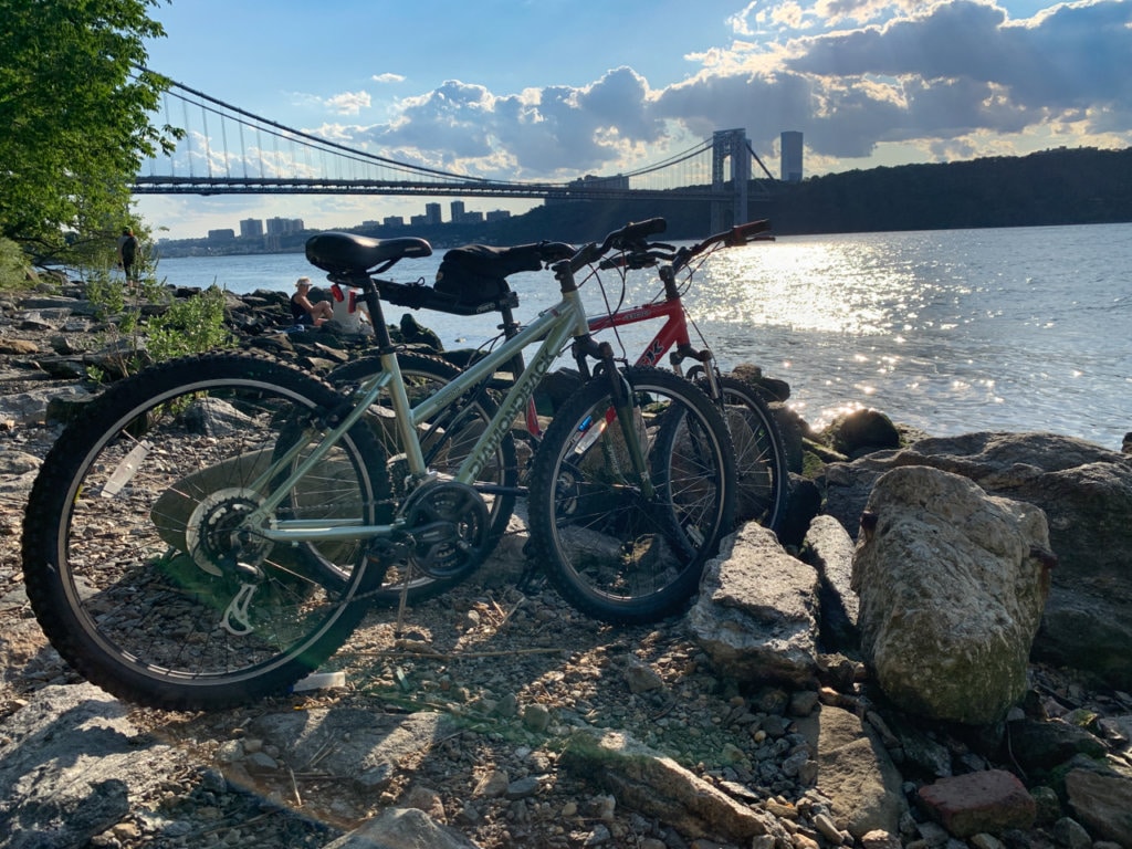 Two bicycles on the shoreline of the Hudson River with the George Washington Bridge in the background.