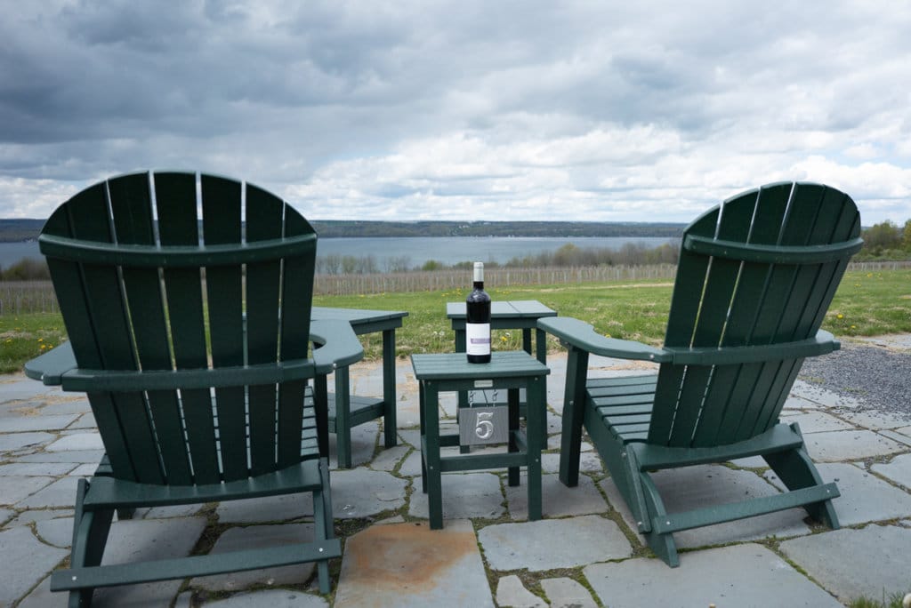 Bottle of wine on a table outside with a green Adirondack chair on either side of the table. 