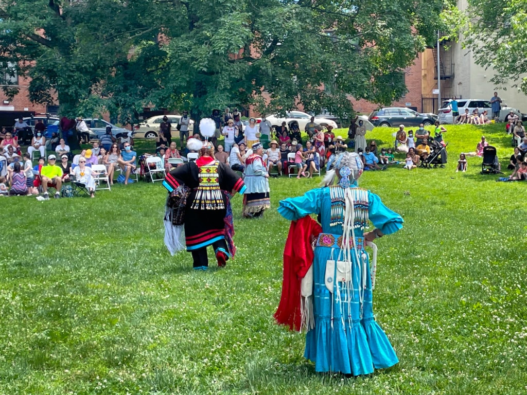 Women dressed in colorful Native American dresses and beads performing a dance in the park in front of an audience.