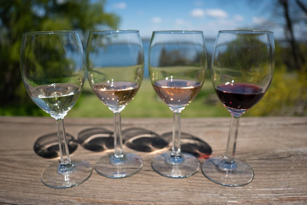 Row of four glasses of wine. Trees and the lake are reflected in the glasses. 