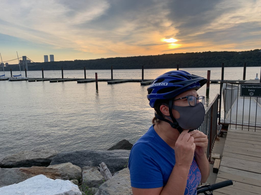 Woman wearing a bicycle helmet and face mask, standing at the shoreline of the Hudson River. 