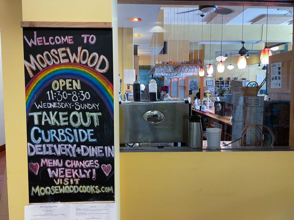 Entrance to a restaurant. Chalkboard sign hanging at entrance says, Welcome to Moosewood, and provides opening hours and additional information.