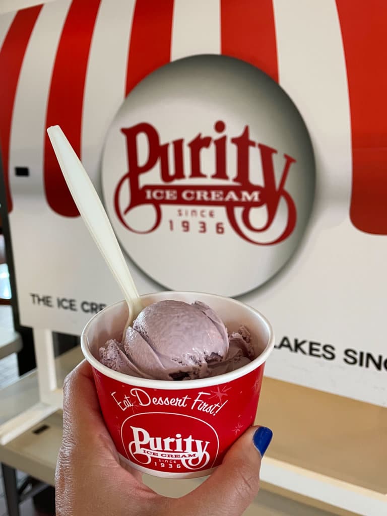 A hand holding a cup of black raspberry ice cream, against a sign that says, Purity Ice Cream, since 1936. 