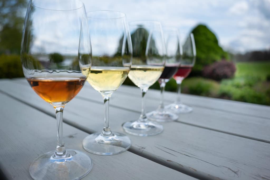A row of 5 glasses of wine, both red and white, sitting on a picnic table outside. 
