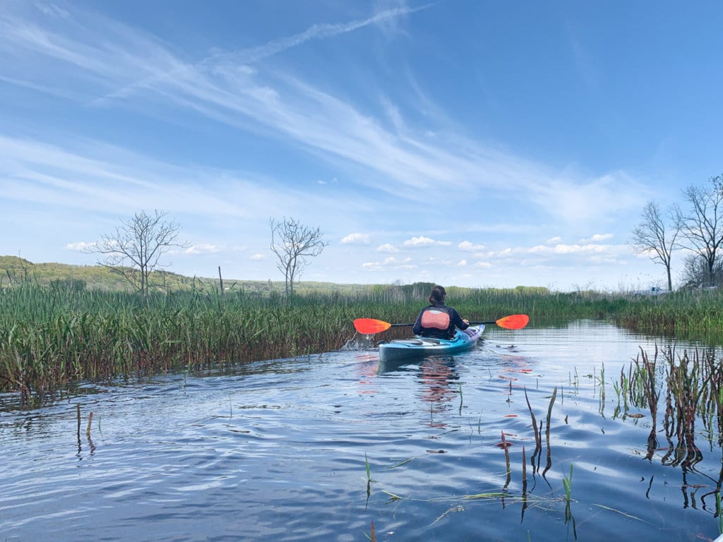 A woman seen from behind paddles in a kayak on in a grassy marsh. 