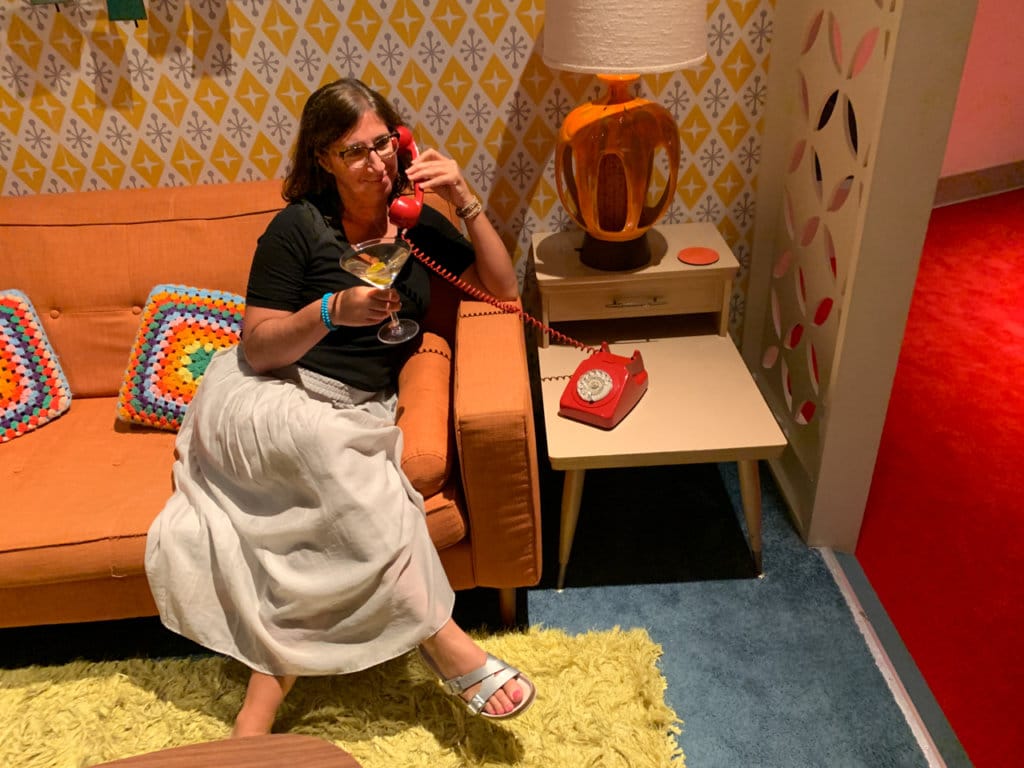 A man sitting on an orange sofa in a living room decorated to reflect the 1960s. 