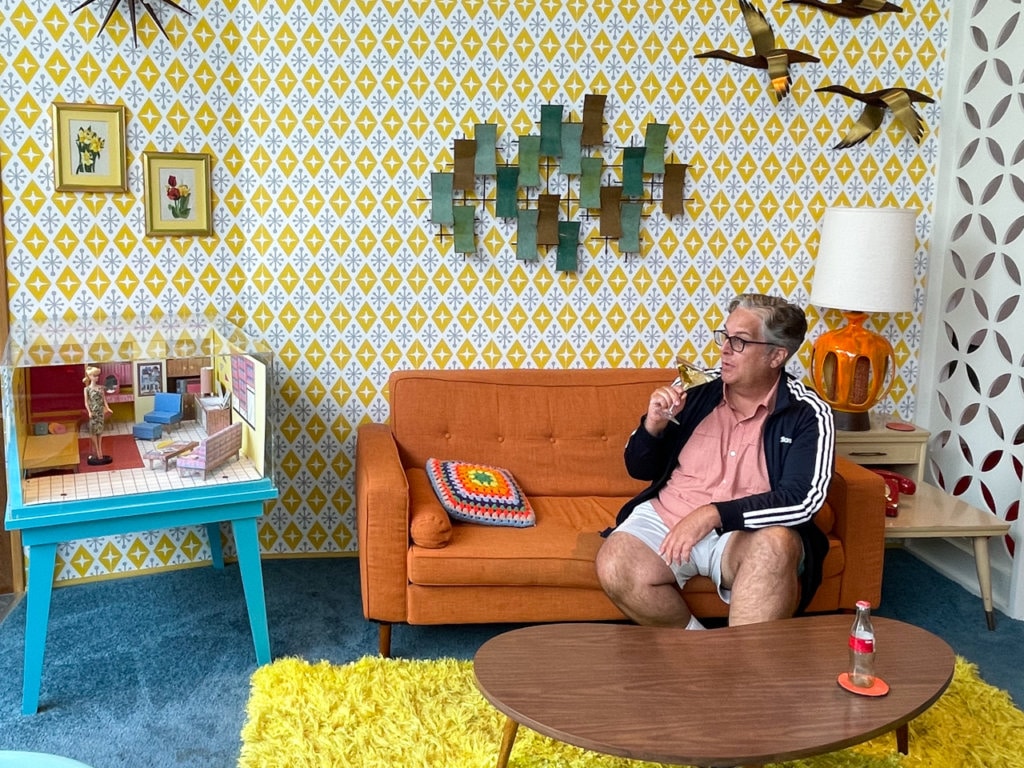 A man sitting on an orange sofa in a living room decorated to reflect the 1960s.