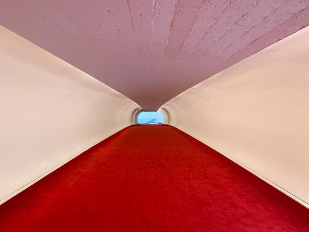Long narrow passageway in airport terminal with bright red carpet, white walls, and rose-colored ceiling. 