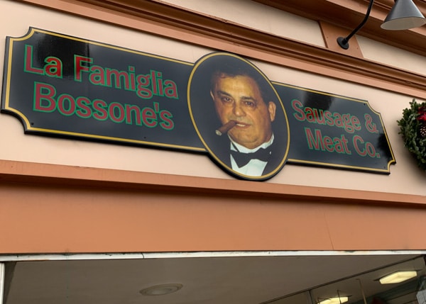 Sign hanging above entrance to a meat market. Sign says La Famiglia Bossone's Sausage & Meat Co. A picture of a man with cigar is also on the sign.