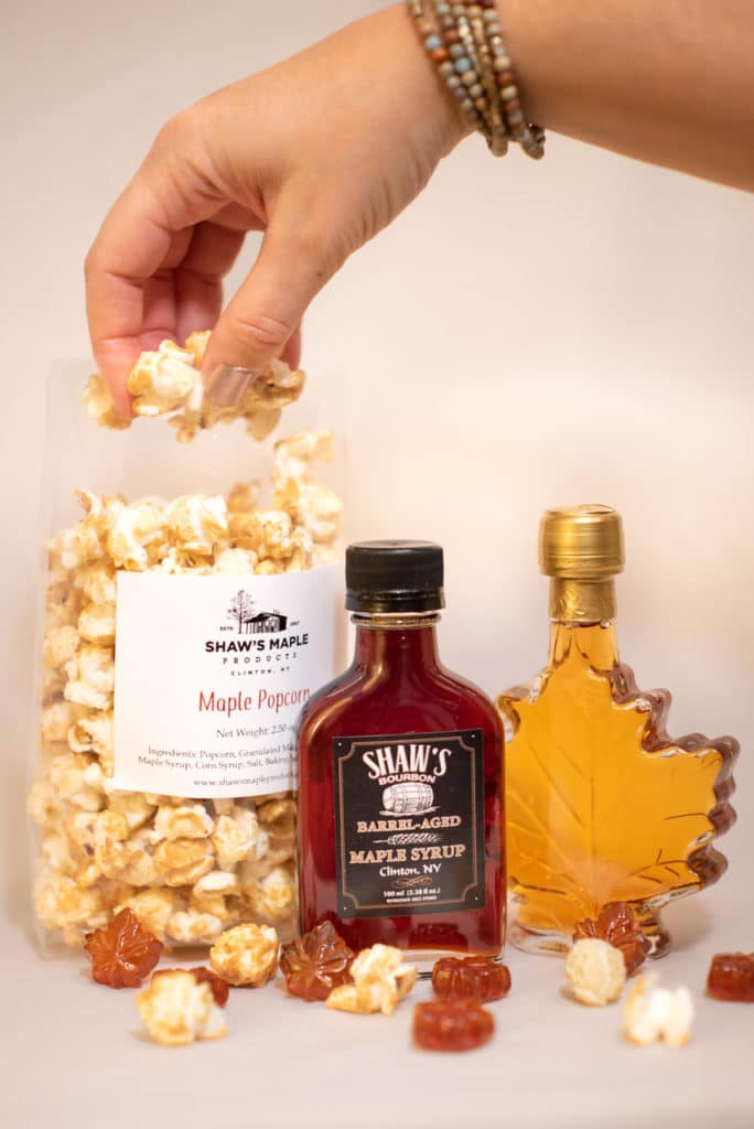 A hand reaching into a bag of maple-flavored popcorn. Two small bottles of maple syrup sit next to the popcorn.